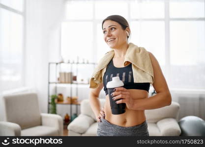 Woman holds water bottle after training. Female person in sportswear, internet sport workout, room interior on background. Woman holds mat, online fitness training