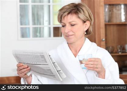 Woman in a bathrobe reading a newspaper and drinking a glass of water