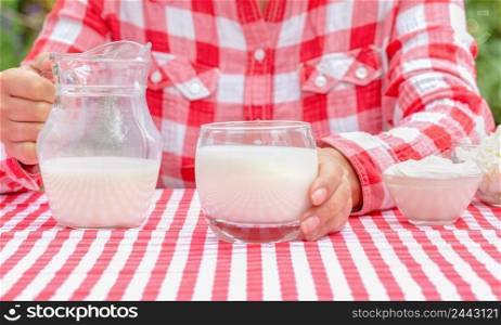 Woman in a checkered red shirt holds out a glass of milk. On the table is a red tablecloth. On the tablecloth is a jug of milk and sour cream. Procurement for an advertising project. Woman in red shirt holds out glass of milk