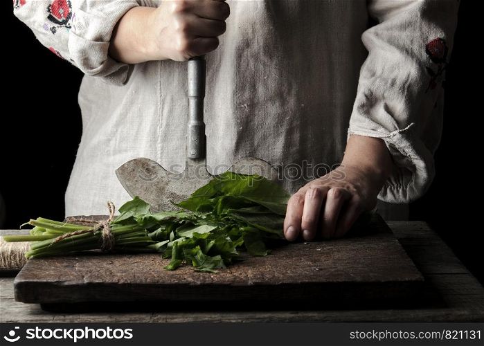 woman in a gray linen dress is cutting green leaves of fresh sorrel on a wooden cutting board with an old metal knife, close up