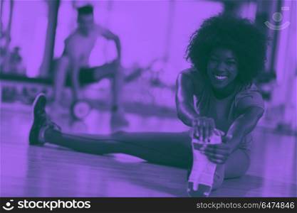 woman in a gym stretching and warming up man in background worki. happy young african american woman in a gym stretching and warming up before workout young mab exercising with dumbbells in background duo tone filter