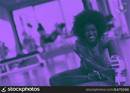 woman in a gym stretching and warming up man in background worki. happy young african american woman in a gym stretching and warming up before workout young mab exercising with dumbbells in background duo tone filter