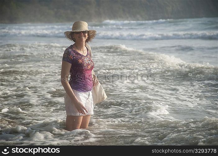 Woman in a hat smiling standing in the ocean surf at knee height