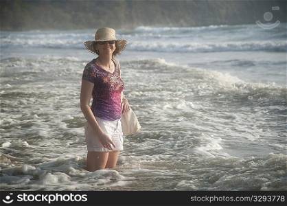 Woman in a hat smiling standing in the ocean surf at knee height