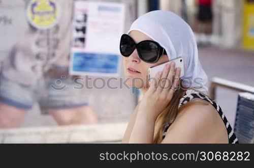 Woman in a headscarf and sunglasses sitting chatting on a mobile phone smiling as she listens to the conversation
