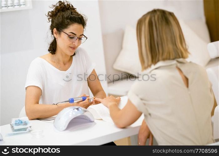 Woman in a nail salon receiving a manicure by a beautician with. Beautician file nails to a customer.. Woman in a nail salon receiving a manicure by a beautician with.