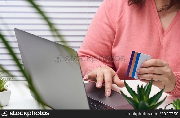 woman in a pink sweater makes purchases online with a laptop, a credit card in hand. Woman sitting at a white table and looking at the monitor