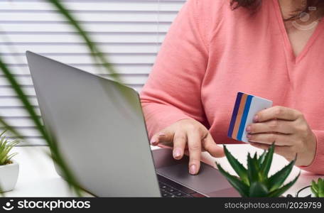 woman in a pink sweater makes purchases online with a laptop, a credit card in hand. Woman sitting at a white table and looking at the monitor