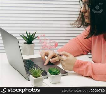 woman in a pink sweater makes purchases online with a laptop, a credit card in hand. A woman sits on a white table and looks at the monitor