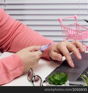woman in a pink sweater makes purchases online with a laptop, a credit card in hand. Woman sitting at a white table