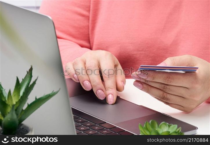woman in a pink sweater makes purchases online with a laptop, a credit card in hand. Woman sitting at white table
