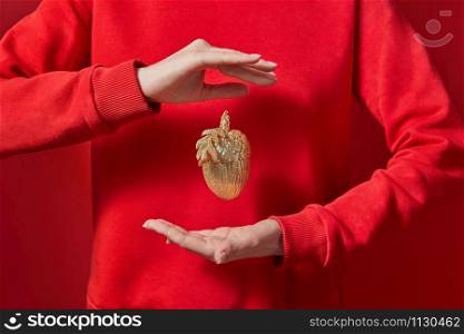 Woman in a red sweatshirt holds beautiful eco Christmas decor golden acorn in hr hands on the same color background with copy space. New Year greeting card.. Hands are hanging golden acorn.