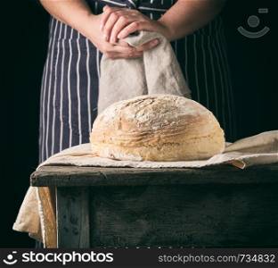 woman in an apron holds a gray linen napkin next to baked round bread on a wooden table, dark background