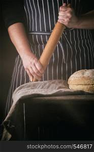 woman in an apron holds a wooden rolling pin next to baked round bread on a wooden table, dark background, sun's rays from the window