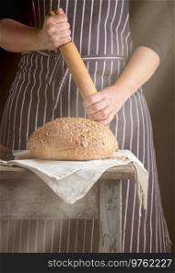 woman in an apron holds a wooden rolling pin next to baked round bread on a wooden table, dark background,  sun’s rays from the window