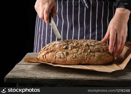 woman in an blue apron with a knife in her hand about to cut round baked bread on a wooden table, dark background