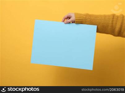 woman in an orange sweater holds a blank sheet of paper on a yellow background. Place for an inscription, advertisement, information