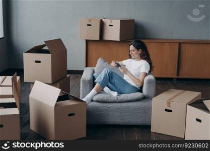 Woman in armchair clicks phone among boxes. European girl having rest in new apartment after relocation. Successful independent woman is moving. Mortgage loan and dream house purchase concept.. Woman in armchair clicks phone among boxes. European girl having rest in apartment after relocation.