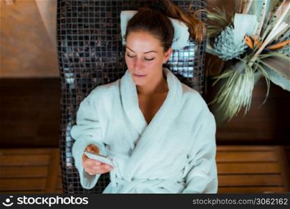 Woman in Bathrobe Lying on a Tepidarium Bed and using a Smart Phone.. Woman Relaxing on a Warm Tepidarium Bed in Spa Center.