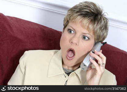 Woman in beige suit speaking on cordless phone. Shocked expression. Blonde hair, green eyes.