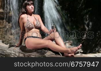woman in bikini drinking fresh water from a glass while sitting by the waterfall