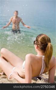 Woman in bikini sunbathing by sea on beach, man in background splashing water