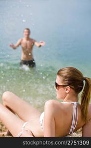 Woman in bikini sunbathing by sea on beach, man in background splashing water