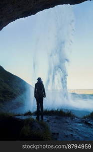 Woman in cave under waterfall landscape photo. Beautiful nature scenery photography with hills on background. Idyllic scene. High quality picture for wallpaper, travel blog, magazine, article. Woman in cave under waterfall landscape photo