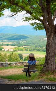 Woman in dress sitting on a bench under the tree overlooking valley and mountains in south France