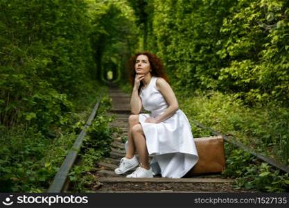 Woman in dress sitting on vintage suitcase on railroad track at tunnel of forest - journey concept
