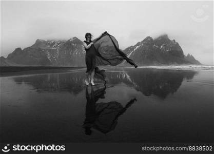 Woman in dress with waving cape on beach monochrome scenic photography. Picture of person with hills on background. High quality wallpaper. Photo concept for ads, travel blog, magazine, article. Woman in dress with waving cape on beach monochrome scenic photography
