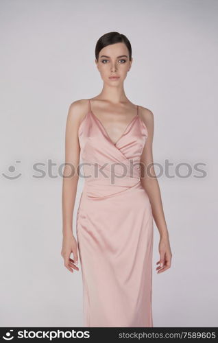 Woman in elegant fashionable dress. Beautiful model pose in studio in pink evening clothes. Classic silk dress. Sexy lady stay on white background. Perfect young woman with classic hairstyle and makeup.