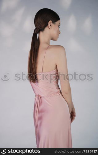 Woman in elegant fashionable dress. Beautiful model pose in studio in pink evening clothes. Classic silk dress. Sexy lady stay on white background. Perfect young woman with classic hairstyle and makeup.