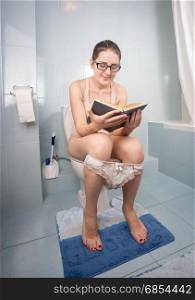 Woman in eyeglasses sitting on toilet and reading book