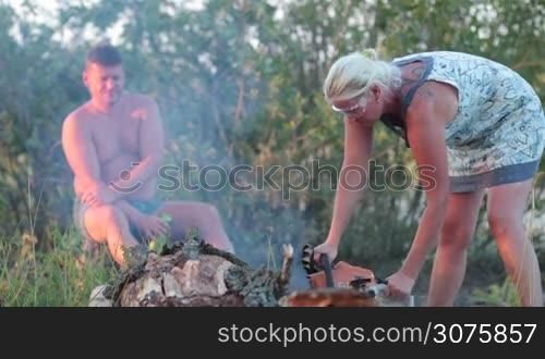 Woman in glasses sawing thick dry tree with chainsaw. Man with can of beer sitting on stump and watching. Happy family picnicking on summer vacation