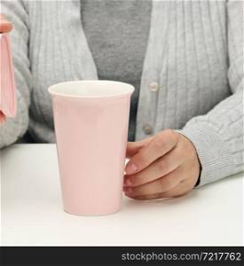 woman in gray clothes holds a pink ceramic cup, woman sits at a white table, start of the day, morning