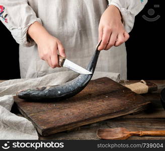 woman in gray linen clothes cleans the fish sea bass scales on a brown wooden board, black background