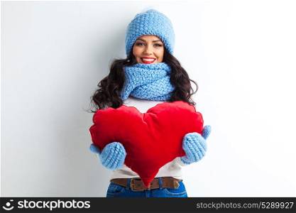 Woman in hat holding heart. Beautiful smiling brunette woman in winter hat and scarf holding a red heart pillow
