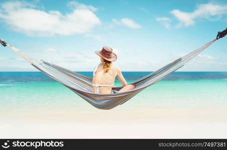 Woman in hat sitting on hammock on the beach and enjoying the beautiful seascape .