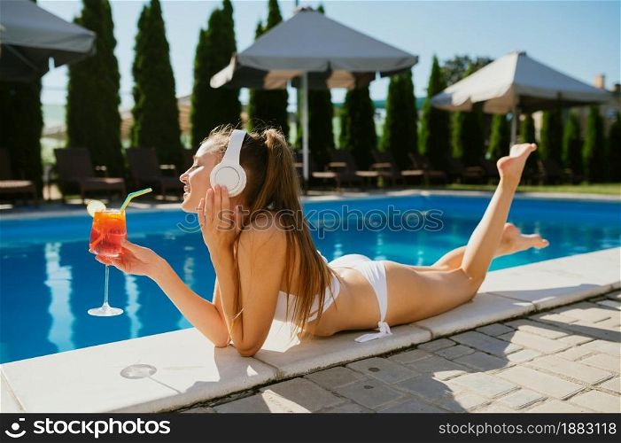Woman in headphones listening to music and drinks coctail at the edge of pool. Happy people having fun on summer vacations, holiday party at the poolside outdoors. Woman in headphones drinks coctail at the pool