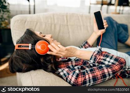 Woman in headphones listening to music on sofa. Pretty lady in earphones relax in the room, sound lover resting on couch. Woman in headphones listening to music on sofa