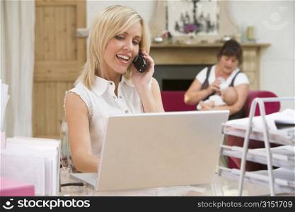 Woman in home office with laptop and telephone with mother and baby in background