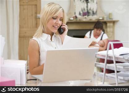 Woman in home office with laptop and telephone with mother and baby in background