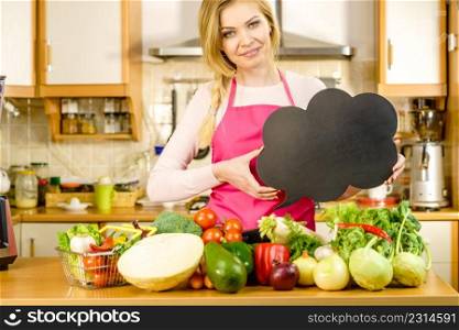 Woman in kitchen wearing apron having many vegetables on table holding speech bubble, thinking balloon black board with copy space. Healty eating, dieting concept.. Woman with speech bubble and vegetables