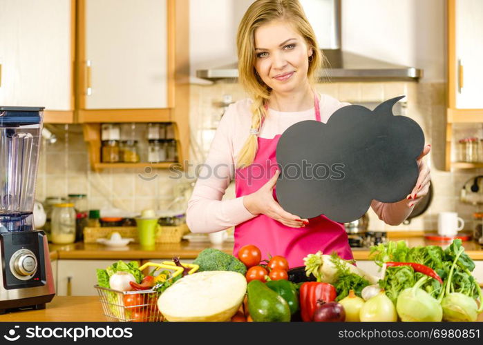 Woman in kitchen wearing apron having many vegetables on table holding speech bubble, thinking balloon black board with copy space. Healty eating, dieting concept.. Woman with speech bubble and vegetables