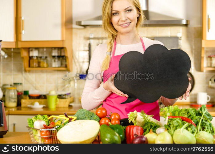 Woman in kitchen wearing apron having many vegetables on table holding speech bubble, thinking balloon black board with copy space. Healty eating, dieting concept.. Woman with speech bubble and vegetables