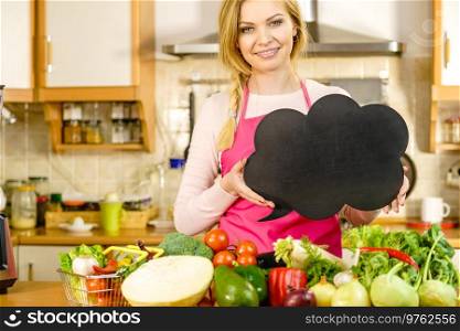 Woman in kitchen wearing apron having many vegetables on table holding speech bubble, thinking balloon black board with copy space. Healty eating, dieting concept.. Woman with speech bubble and vegetables