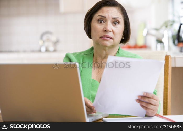 Woman in kitchen with laptop and paperwork looking worried