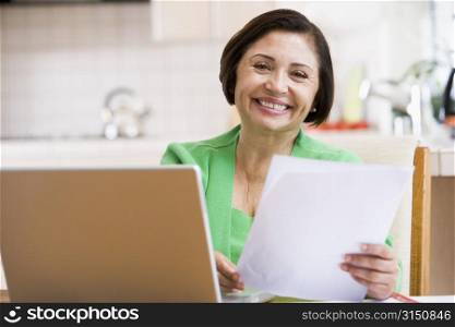 Woman in kitchen with laptop and paperwork smiling