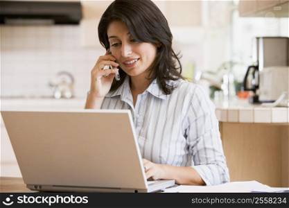 Woman in kitchen with laptop using cellular phone smiling